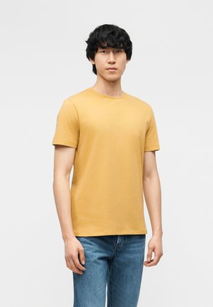 Jeune homme aux cheveux noirs debout portant un t-shirt à manches courtes jaune moutarde et un jean bleu sur un fond gris clair uni.