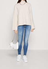 Pull beige à col roulé avec manches larges, associé à un jean skinny bleu. Sac à main blanc avec bandoulière chaîne argentée et baskets blanches.