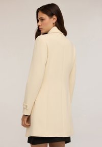 Manteau couleur crème avec une coupe ajustée, doté d'un col cranté et de poignets boutonnés. Tissu lisse avec une silhouette élégante.