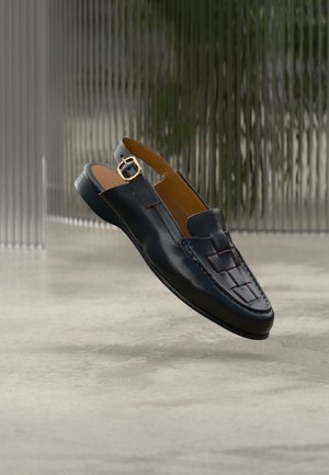 Melns ādas slingbeka stila loafer apavs ar pinumu priekšpusē, zelta sprādzīti un plakanu zoli, attēlots uz pelēka, teksturēta fona.