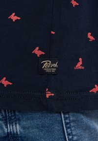 T-paita tummansinisellä pohjalla, jossa on pieniä punaisia logoja. Sivusaumassa on kudottu musta etiketti, jossa on kultateksti. Silkkinen puuvillapinta.