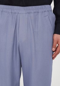 Pantalon bleu clair à taille élastique avec poches latérales, porté avec une chemise noire, montrant un gros plan sur le bas du torse et la main droite.
