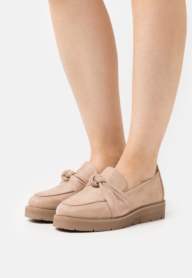 Mocassins beige en daim sans lacets avec un détail noué, portés pieds nus sur un fond blanc.