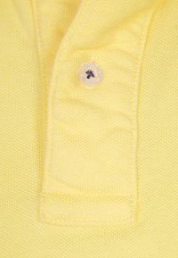 Žlutá polo košile zblízka s texturovanou látkou, knoflíkem s tmavě modrou nití a složeným límcem.