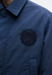 Marineblauwe jas van gestructureerde stof, met een ronde geborduurde logo met "BOSS" en "EST. 1971" op de linkerborst.
