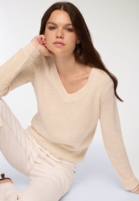 Beżowy sweter z dekoltem w serek, o prążkowanej strukturze i długich rękawach, zestawiony z jasnymi, dopasowanymi spodniami.