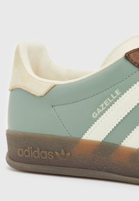Primer plano de una zapatilla Adidas Gazelle verde con rayas blancas, lengüeta beige en el talón y suela marrón texturizada.