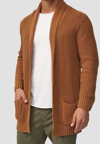 Sweter kardigan w ciepłym odcieniu brązu, z kołnierzem szalowym, otwartym przodem i dwoma przednimi kieszeniami, posiadający prążkowane mankiety i dół.