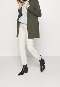 Persona que lleva un abrigo color verde oliva, sudadera gris clara, pantalones blanco roto y botines negros de tacón, de pie contra un fondo claro y liso.