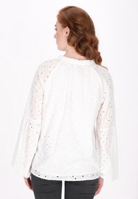 Weiße, bestickte Bluse mit Rüschenausschnitt und Glockenärmeln; verfügt über Blumenmuster und Durchbrüche.
