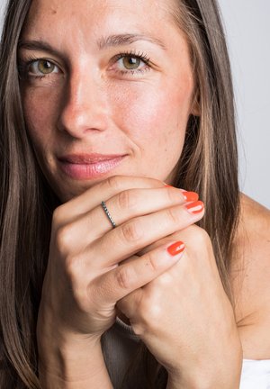 Primo piano delle mani di una donna con smalto arancione, che tiene il mento, indossando un sottile anello d'argento, con pelle liscia e lunghi capelli castani.