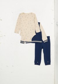 Beige langermet bodysuit med mønsterdesign og myke marineblå bukser med hvite striper og trykknapper ved mansjettene. Henger mot en hvit vegg.