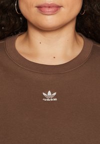 Närbild av en person som bär en brun Adidas-tröja med den vita logotypen centrerad under halsringningen och guldörhängen synliga.
