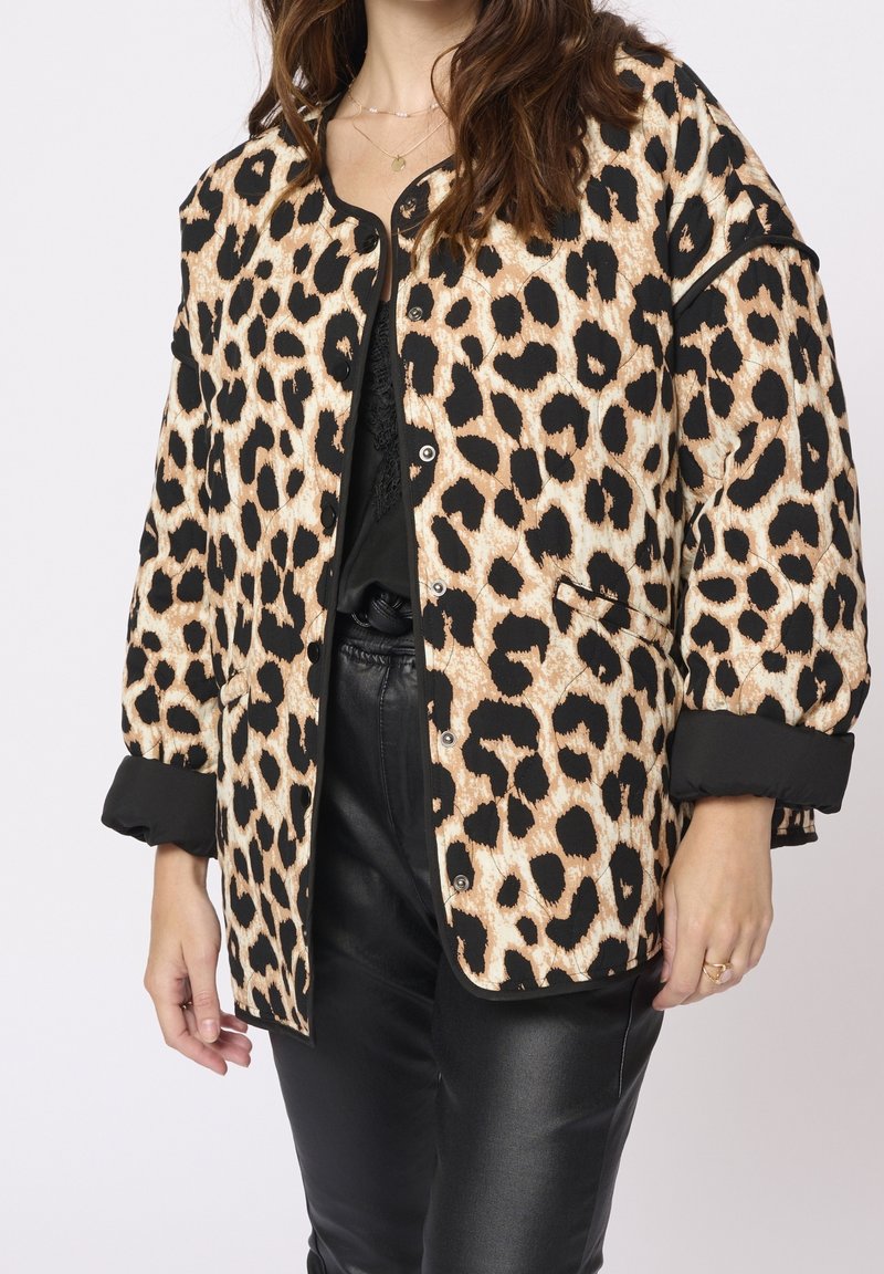 Femme portant une veste boutonnée à imprimé léopard par-dessus un top noir et un pantalon en cuir noir, avec des colliers superposés et une bague à une main.