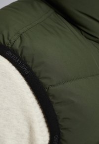 Green puffer vest with a textured cream underlayer. Features black elastic banding at the armhole, showcasing stitching details.