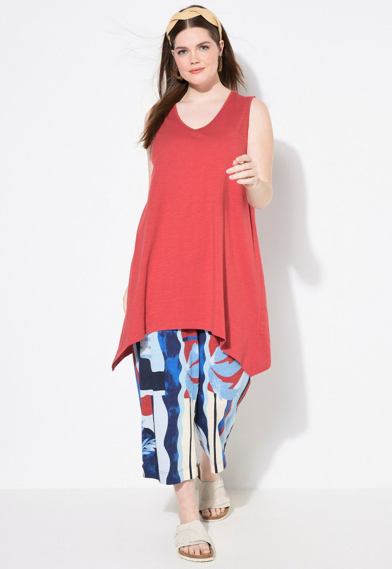 Tunica rossa senza maniche con scollo a V, abbinata a pantaloni larghi blu e con fantasia. Modella che indossa sandali beige e una fascia per capelli.