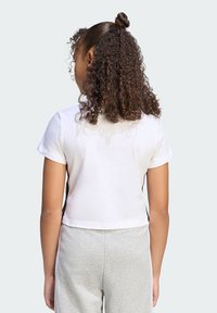 Biela skrátená tričko s krátkymi rukávmi; s čiernymi bočnými akcentmi. Model nosí sivé tepláky. Kučeravé vlasy sú upravené do drdolu.