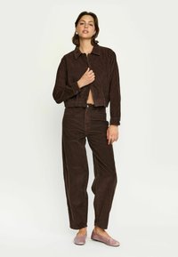 Jachetă scurtă din corduroy maro, asortată cu pantaloni largi care au un finisaj texturat și nervuri verticale subtile. Încălțămintea este formată din balerini de culoare roz deschis.