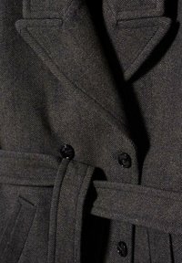 Manteau en laine chevrons gris foncé avec une taille ceinturée, de larges revers, et des boutons noirs. Le tissu texturé ajoute un intérêt visuel.