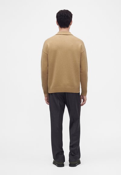 Homme debout de dos portant un pull beige, un pantalon noir et des chaussures noires sur un fond blanc uni.
