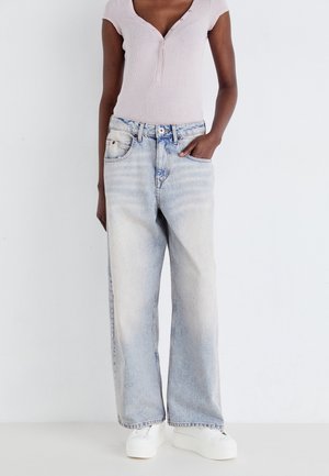 Persoon die een lichtroze top met korte mouwen draagt, lichtblauwe wijde jeans met vervaging en witte platform sneakers, één hand in de zak.