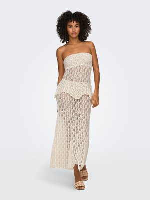 Femme aux cheveux bouclés portant une robe bustier en dentelle beige et des talons ouverts tissés beiges, debout devant un fond clair uni.