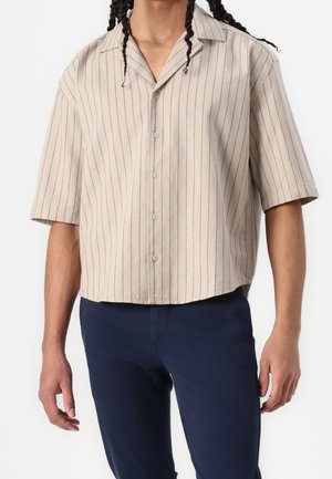 Man draagt een beige overhemd met korte mouwen en dunne verticale strepen, gecombineerd met een donkerblauwe broek, staand tegen een witte achtergrond.