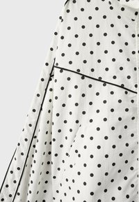 White fabric with black polka dots featuring a diagonal seam and a pocket, outlined with thin black piping.