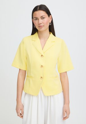 Femme portant un blazer jaune à manches courtes boutonné et une jupe blanche, debout devant un fond clair uni.