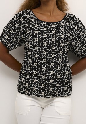 Haut noir avec un motif texturé de cercles blancs entrelacés, manches courtes bouffantes et encolure carrée. Associé à un pantalon blanc.