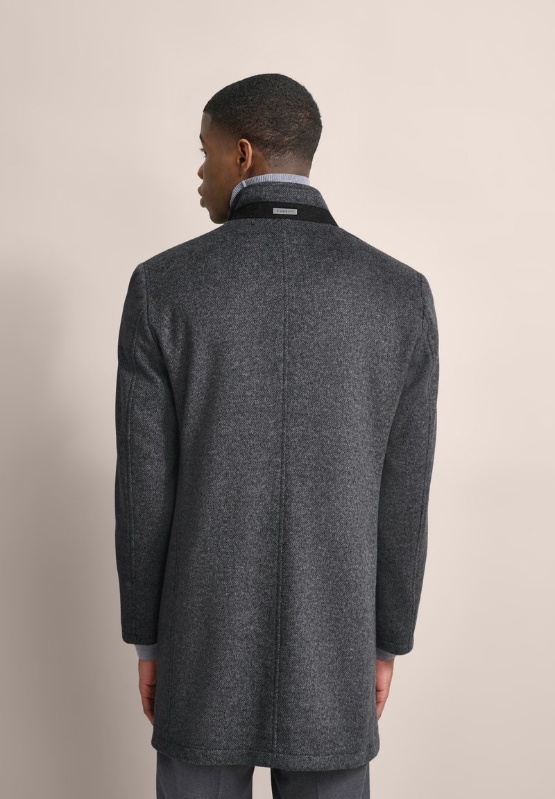 Cappotto grigio in lana con un motivo a spina di pesce, dotato di un colletto nero e maniche lunghe, visto da dietro contro uno sfondo neutro.