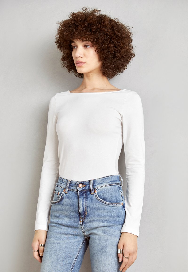 Bodysuit blanc à manches longues avec un décolleté large, associé à un jean bleu taille haute. Tissu lisse, design ajusté et accents minimalistes.