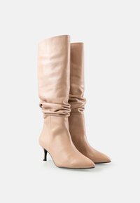 Cizme din piele bej, cu design lejer, până la genunchi, cu vârf ascuțit și toc mic, pe un fundal alb.