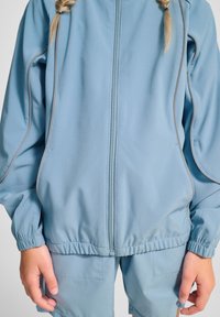 Ljusblå jacka i texturerat tyg, med dragkedja, elastiska ärmmuddar och kontrasterande grå sömmar, tillsammans med matchande shorts.