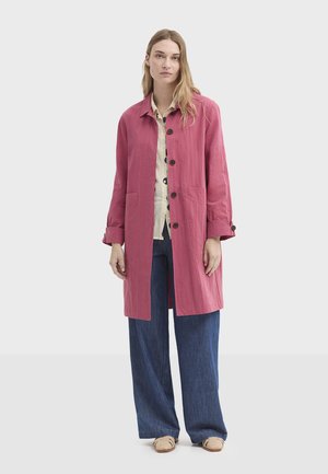 Rosa Leinenmantel mit Kragen, langen Ärmeln, Knopfleiste und zwei Fronttaschen, kombiniert mit blauen Hosen mit weitem Bein und leichten Schuhen.