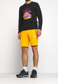 Mann trägt ein schwarzes Langarmshirt mit buntem Grafikdesign, leuchtend orangefarbene Shorts von The North Face, weiße Socken und schwarze Sneakers.