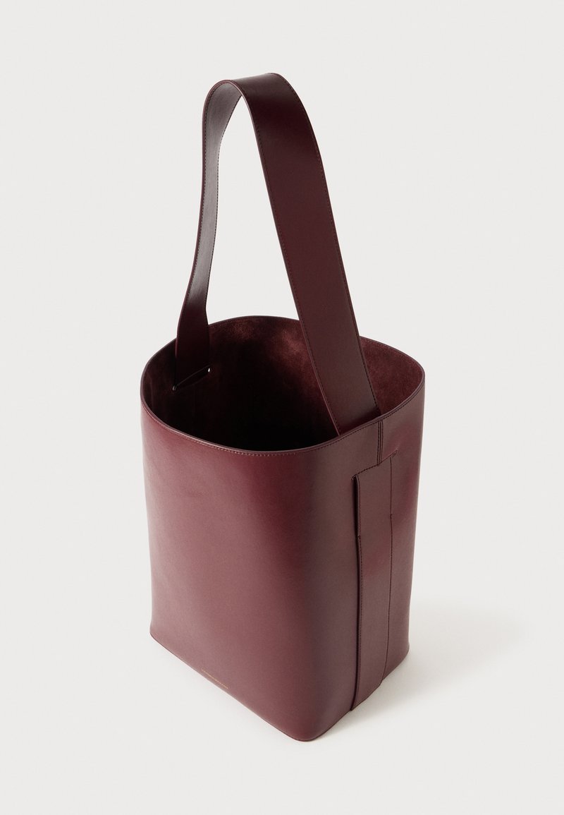 Sac fourre-tout en cuir bordeaux avec une texture lisse, de larges sangles et un dessus ouvert, arborant un design minimaliste et des bords sans couture.