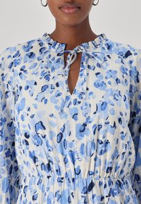 Leichtes Kleid aus Stoff mit einem blauen Blumenmuster auf weißem Hintergrund. Rüschenkragen, Schlitzdetail und geraffte Taille.