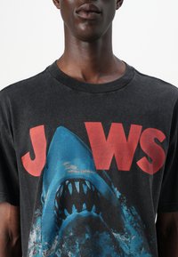 Svart bomull t-skjorte med et blått hai-motiv og stor rød tekst som står "JAWS," med rund hals og avslappet passform.