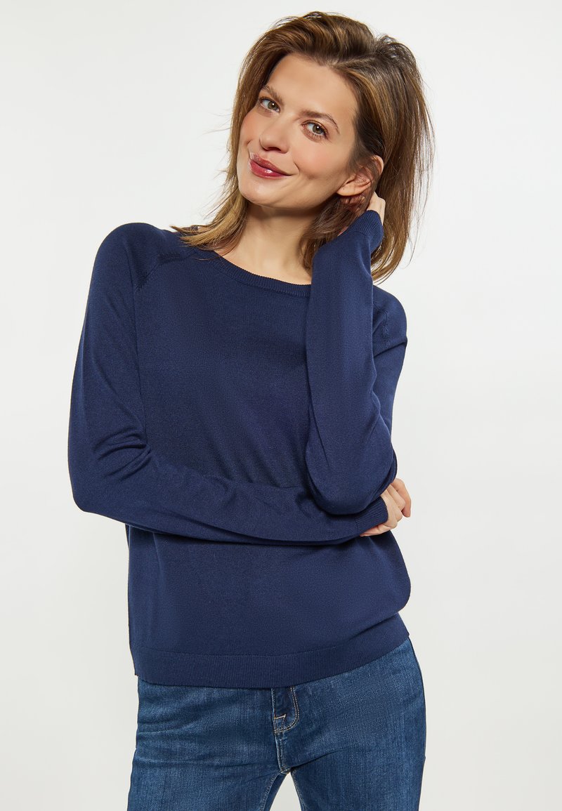 Dunkelblauer Strickpullover mit langen Ärmeln und rundem Ausschnitt, kombiniert mit dunkelblauen Jeans. Der Stoff wirkt weich und strukturiert.