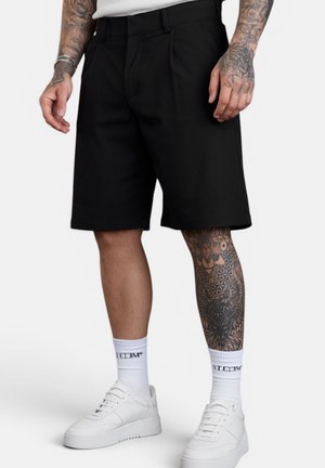 Mann trägt schwarze knielange Shorts, weiße Socken und weiße Sneakers und zeigt vollflächige Bein- und Arm-Tattoos vor einem schlichten Hintergrund.