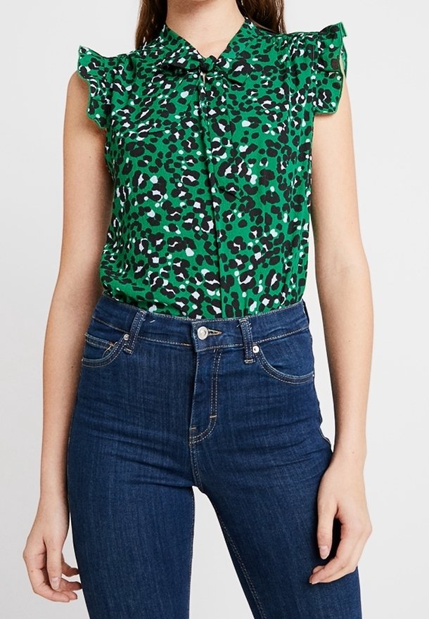 Zelená bezrukávová blůza s leopardím vzorem, se zavazovací přední částí, texturovanými volány na ramenou a lehkým materiálem. Vzhled doplněn modrými džíny.