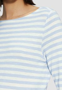 Žena nosící světle modré a bílé tričko s dlouhými rukávy a vodorovnými pruhy, s výstřihem na šířku lodi, oříznuté u ramen a horní části hrudníku.