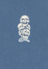 Pět naskládaných lebek s otevřenými ústy a prázdnými očními důlky na modrém pozadí, s stylizovaným textem dole.