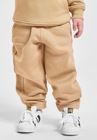 Kind trägt beige Sweatpants und einen passenden Sweatshirt mit weißen Adidas Sneakers mit schwarzen Streifen, stehend auf weißem Hintergrund.