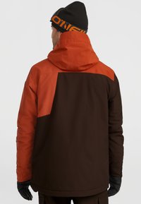 Geïsoleerde jack met een oranje bovenste deel en een donkerbruin onderste deel. Voorzien van een capuchon, textuurstof en zwarte handschoenen.