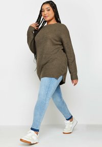 Olivgrüner gestrickter Pullover mit langen Ärmeln, kombiniert mit hellblauen Skinny-Jeans und weißen Sneakers mit braunen Sohlen. Verfügt über ein seitlich geschlitztes Design.