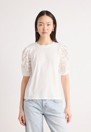 Weißes Baumwoll-T-Shirt mit puffigen Spitzenärmeln, rundem Halsausschnitt und lässiger Passform. Getragen mit hellblauen, verwaschenen Jeans.