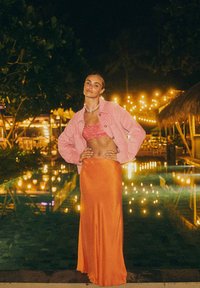 Modelka ubrana w różową materiałową kurtkę, wzorzysty różowy top bikini i pomarańczową, zwiewną maxi spódnicę, stojąca przy basenie z lampkami w tle.