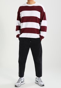 Chemise à manches longues rayée bordeaux et blanche, associée à un pantalon noir ajusté et des chaussures de sport grises. Le tissu semble doux et décontracté.